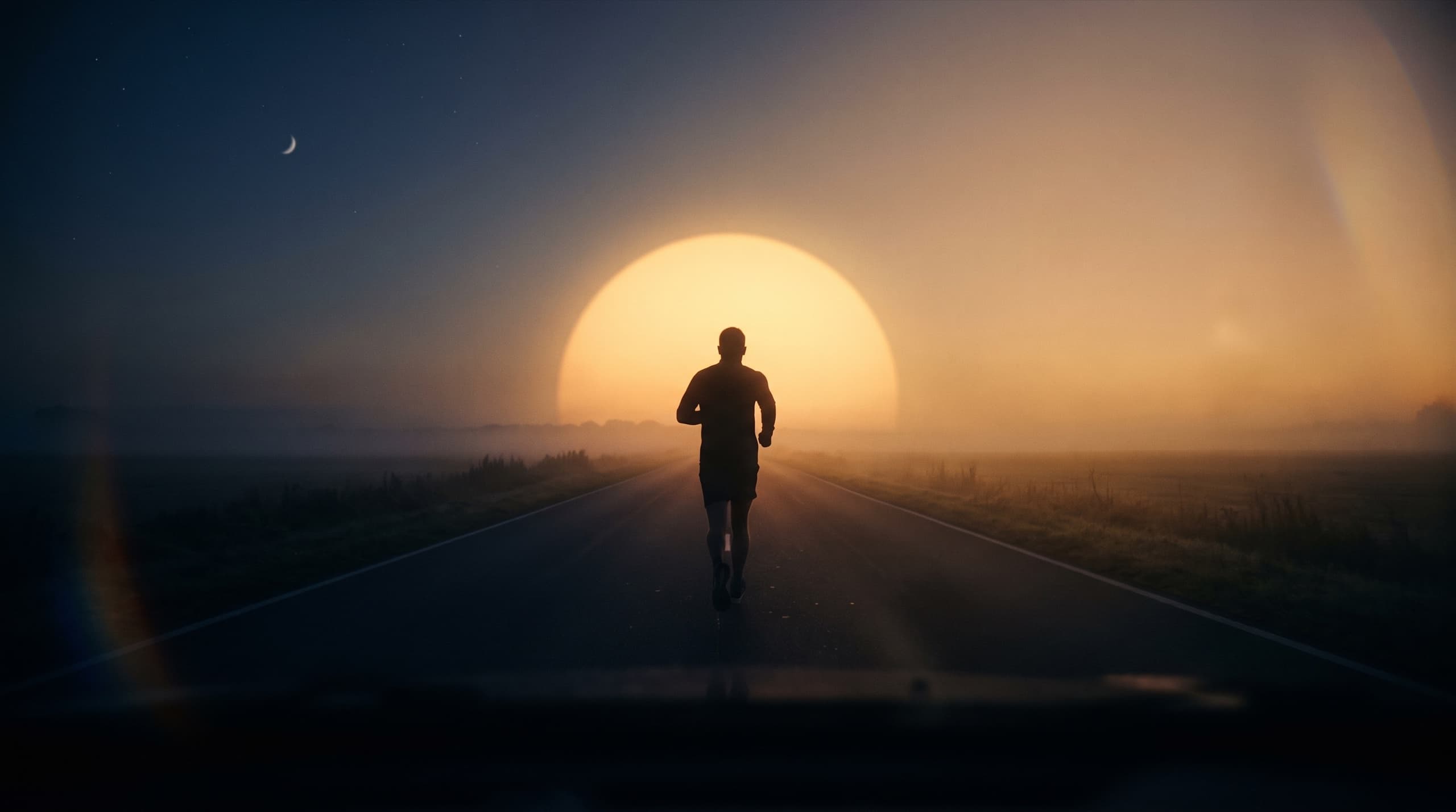 A lone runner on an empty road at dawn, silhouetted against a huge low sun, the moon still visible in the dark half of the sky