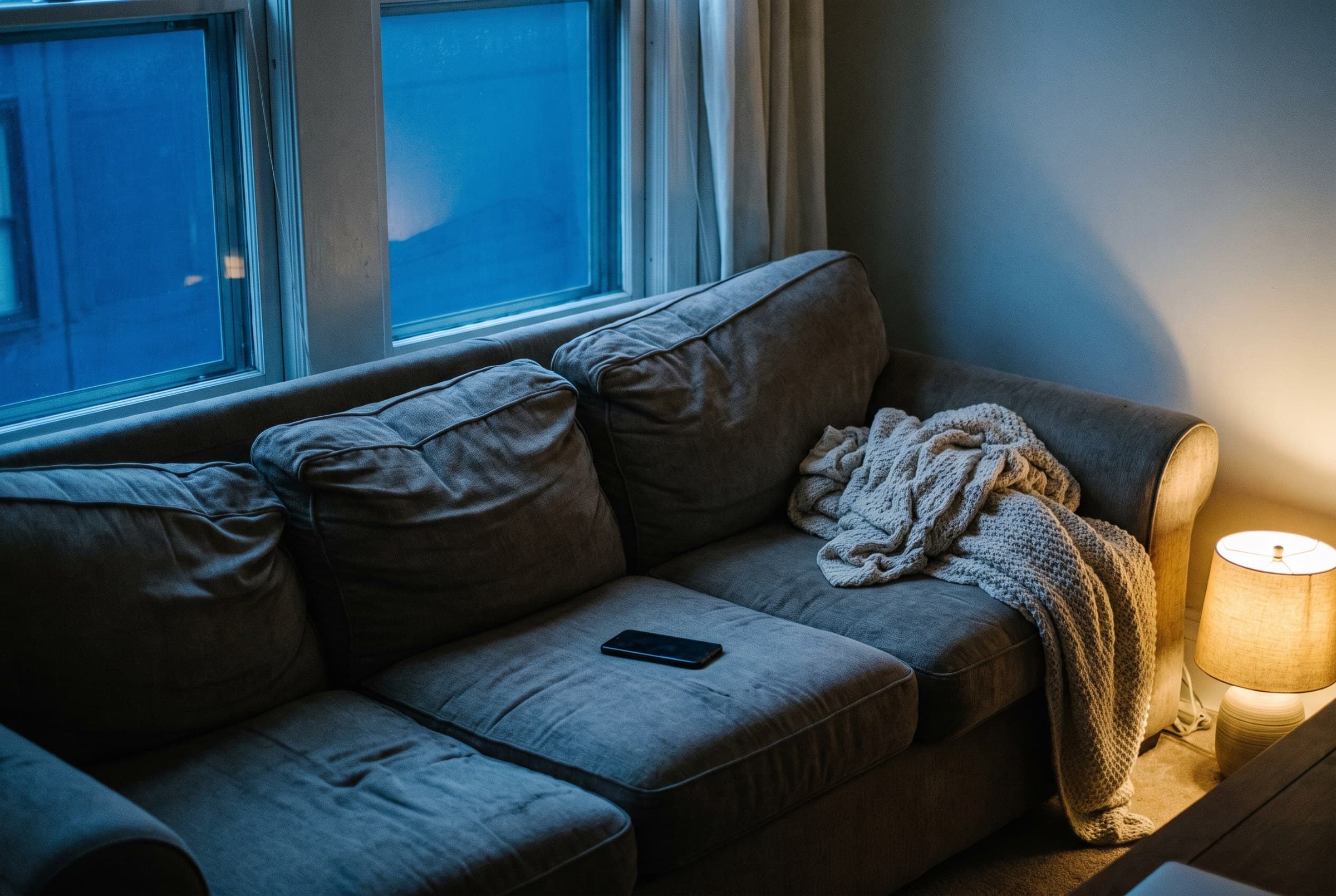 A sagging grey couch at midnight with a dark switched-off phone face-down on a dented cushion, a knitted throw bunched at one end and a small lamp glowing in the corner